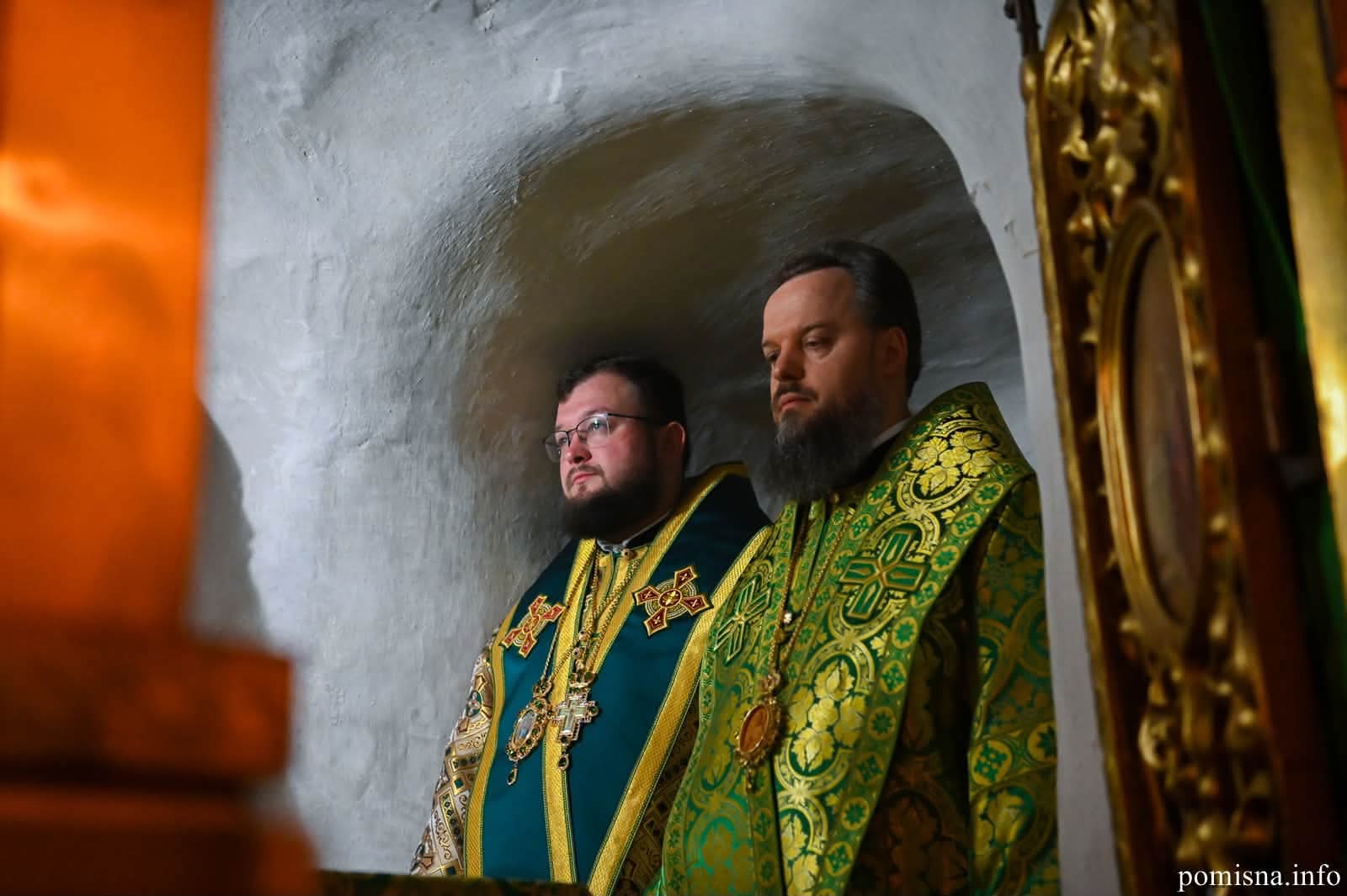 Двоє священнослужителів у зелених облаченнях з панагіями стоять у побіленій ніші підземного храму Ближніх печер