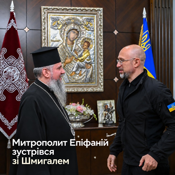 Митрополит Епіфаній у чорному клобуку розмовляє з Денисом Шмигалем у темній куртці з шевроном прапора України; позаду ікона Богородиці з Дитям у срібному окладі, хоругва і прапор України; напис «Митрополит Епіфаній зустрівся зі Шмигалем»